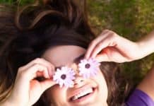 Τί λέει η θετική ψυχολογία για την αισιοδοξία; a happy woman lying in the mown and she cover hers eyes with pink flowers