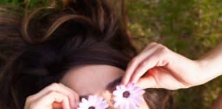 Τί λέει η θετική ψυχολογία για την αισιοδοξία; a happy woman lying in the mown and she cover hers eyes with pink flowers