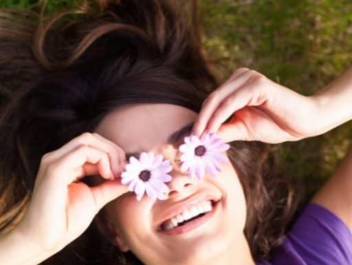 Τί λέει η θετική ψυχολογία για την αισιοδοξία; a happy woman lying in the mown and she cover hers eyes with pink flowers